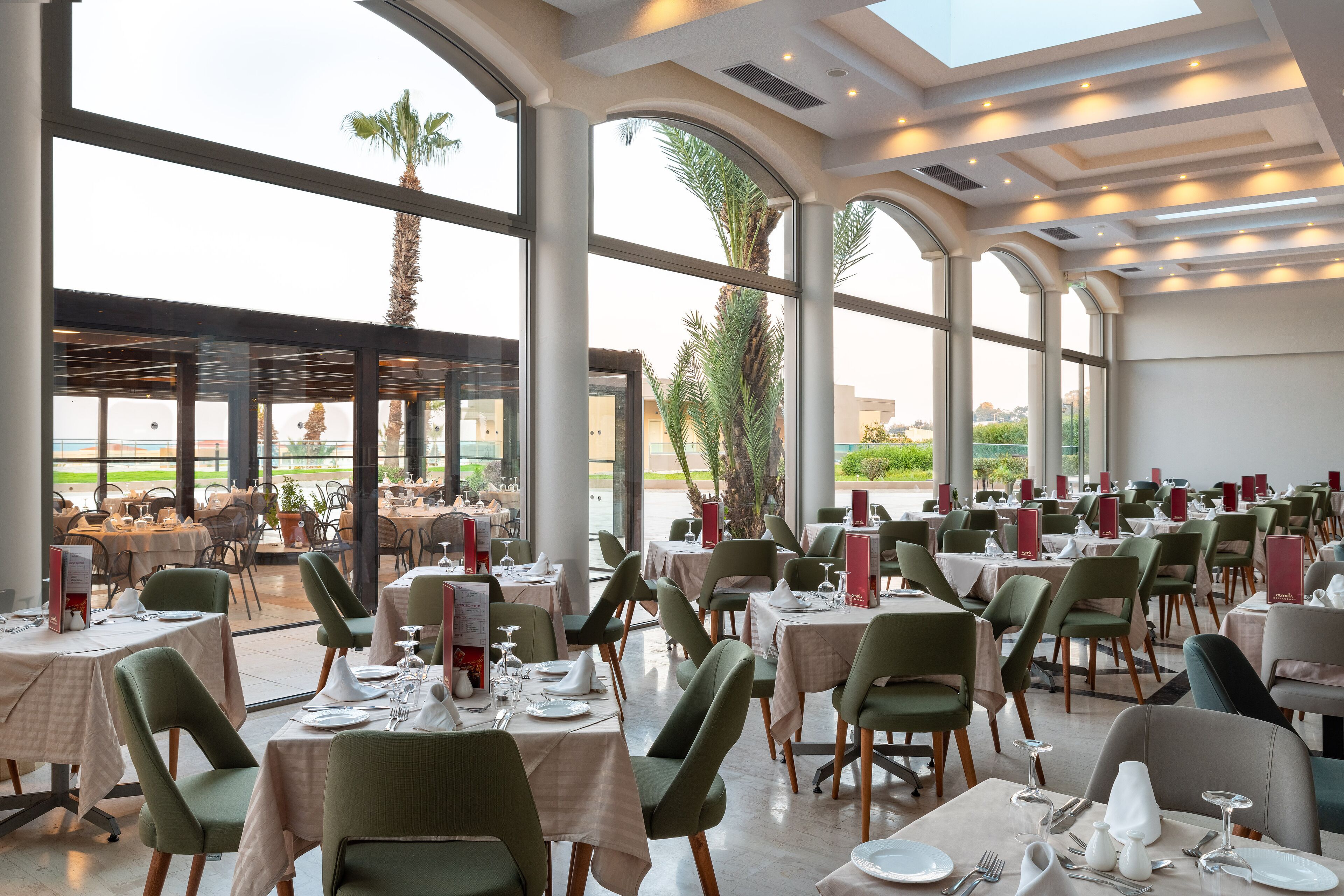 A spacious restaurant with high ceilings, large windows, and a view of palm trees. Tables are set with white tablecloths and green chairs.