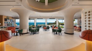 Een ruime binnenruimte met een hoge plafond, grote ramen met uitzicht op de oceaan, en diverse zithoekjes, inclusief tafels en stoelen.