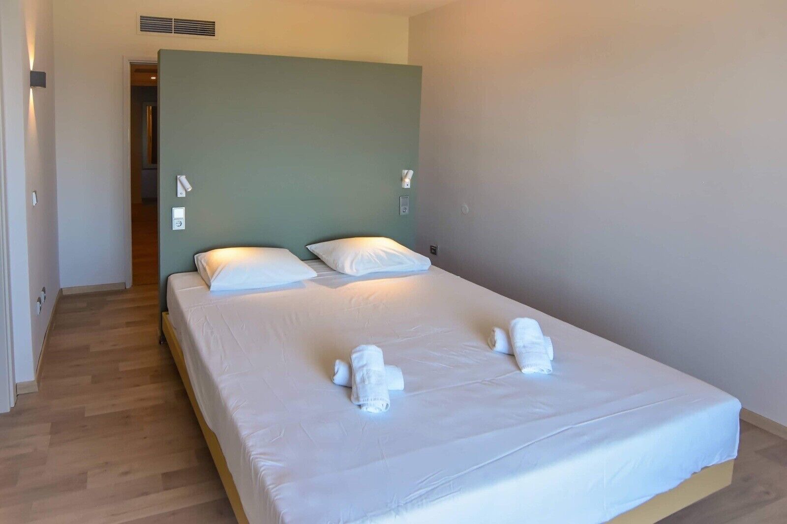 A neatly made bed with white linens and pillows, a wooden floor, and a green accent wall.