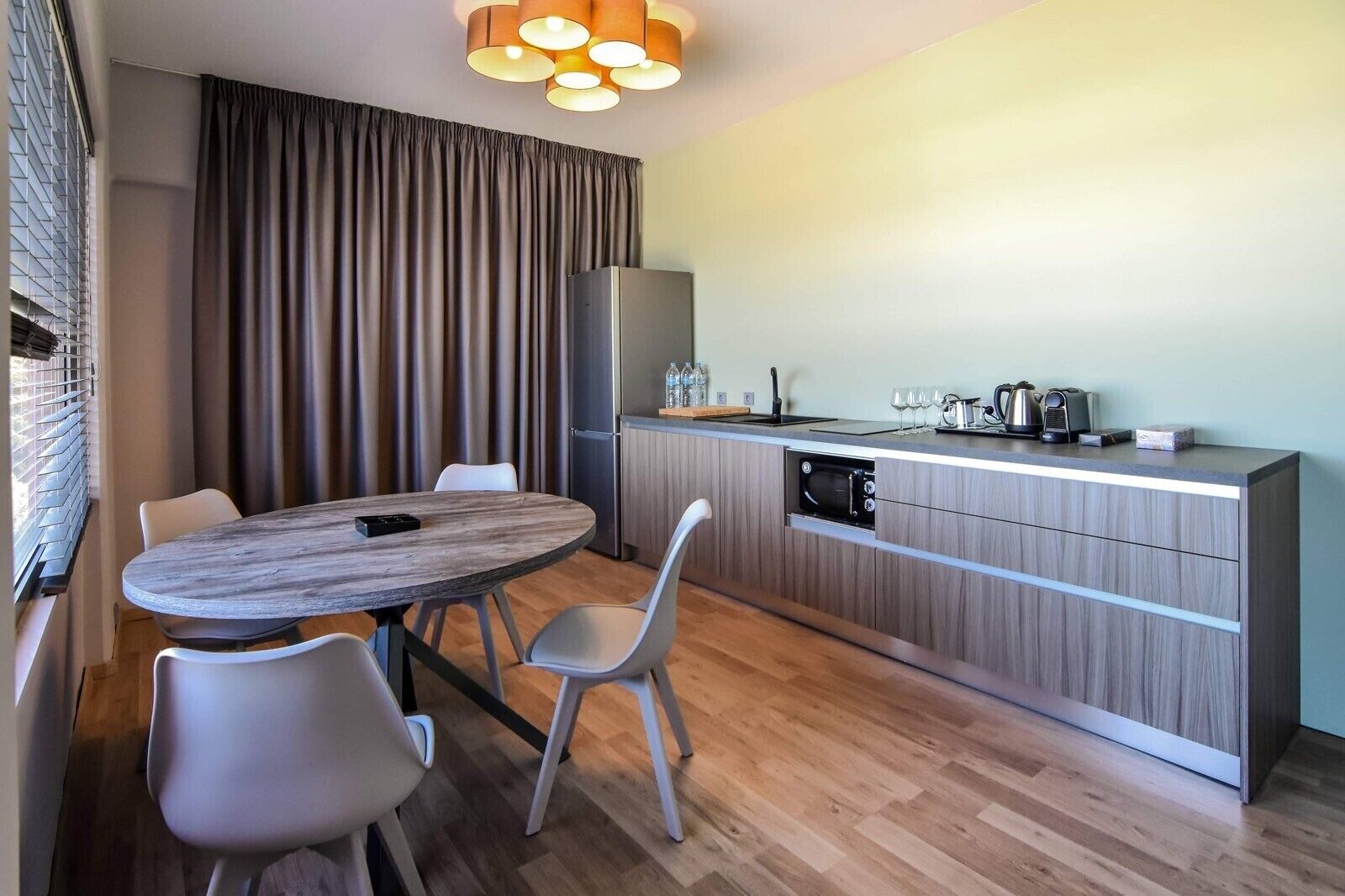 A modern kitchen with a built-in oven, a dining table with chairs, and a large window with blinds.