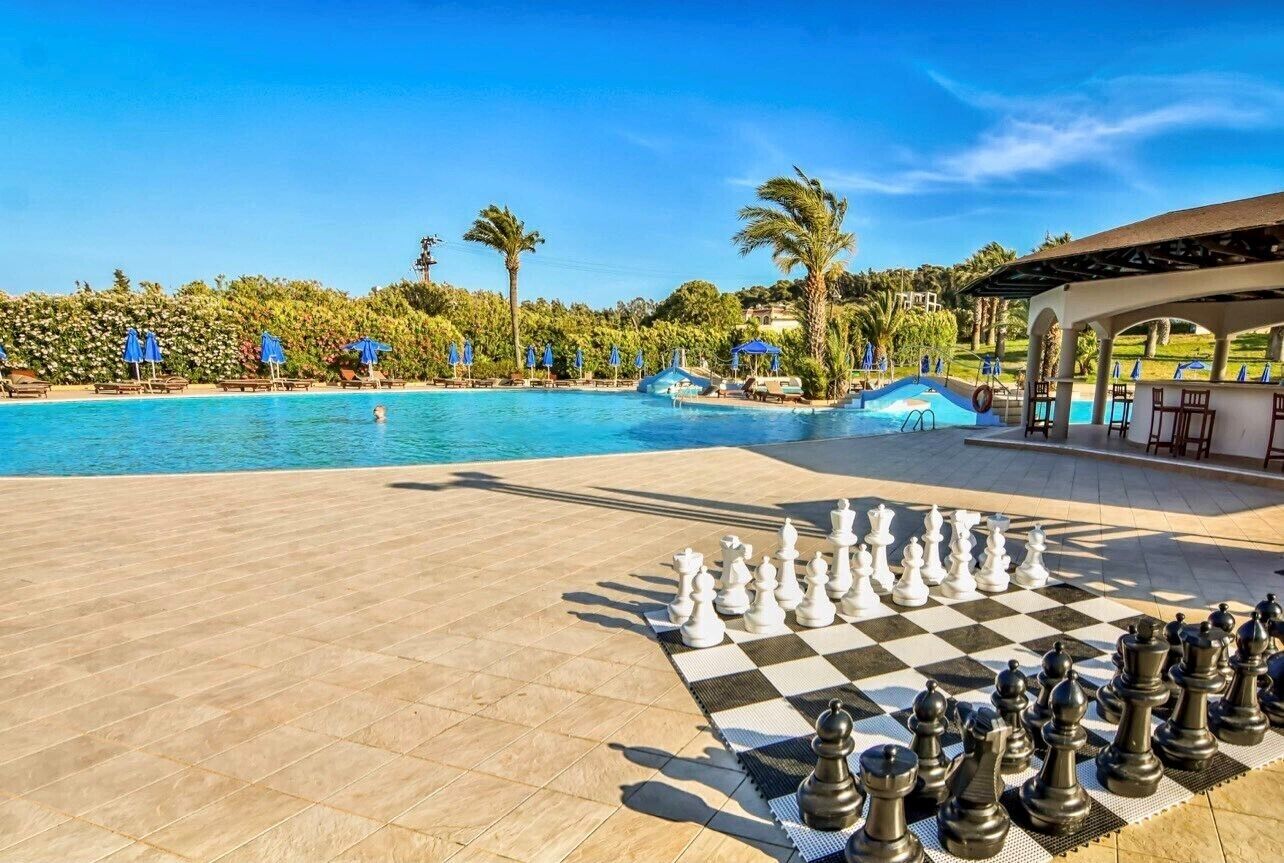 Un tablero de ajedrez junto a la piscina, con un tapete a cuadros, rodeado de sillas de descanso y sombrillas, y una vista despejada de la piscina y las palmeras al fondo.