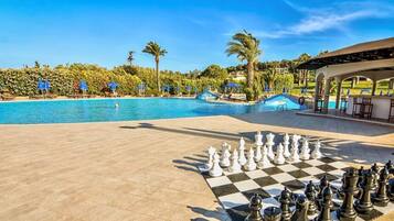 Un tablero de ajedrez junto a la piscina, con un tapete a cuadros, rodeado de sillas de descanso y sombrillas, y una vista despejada de la piscina y las palmeras al fondo.