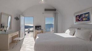 A modern bedroom with a large bed, a desk, a chair, and a television. There is a large window with a view of the outdoors. - Villa Renos (Santorini)