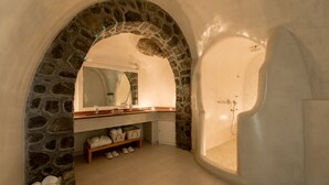 A bathroom with a stone archway, a bathtub, a sink with a mirror, and a shelf with towels. - Villa Renos (Santorini)