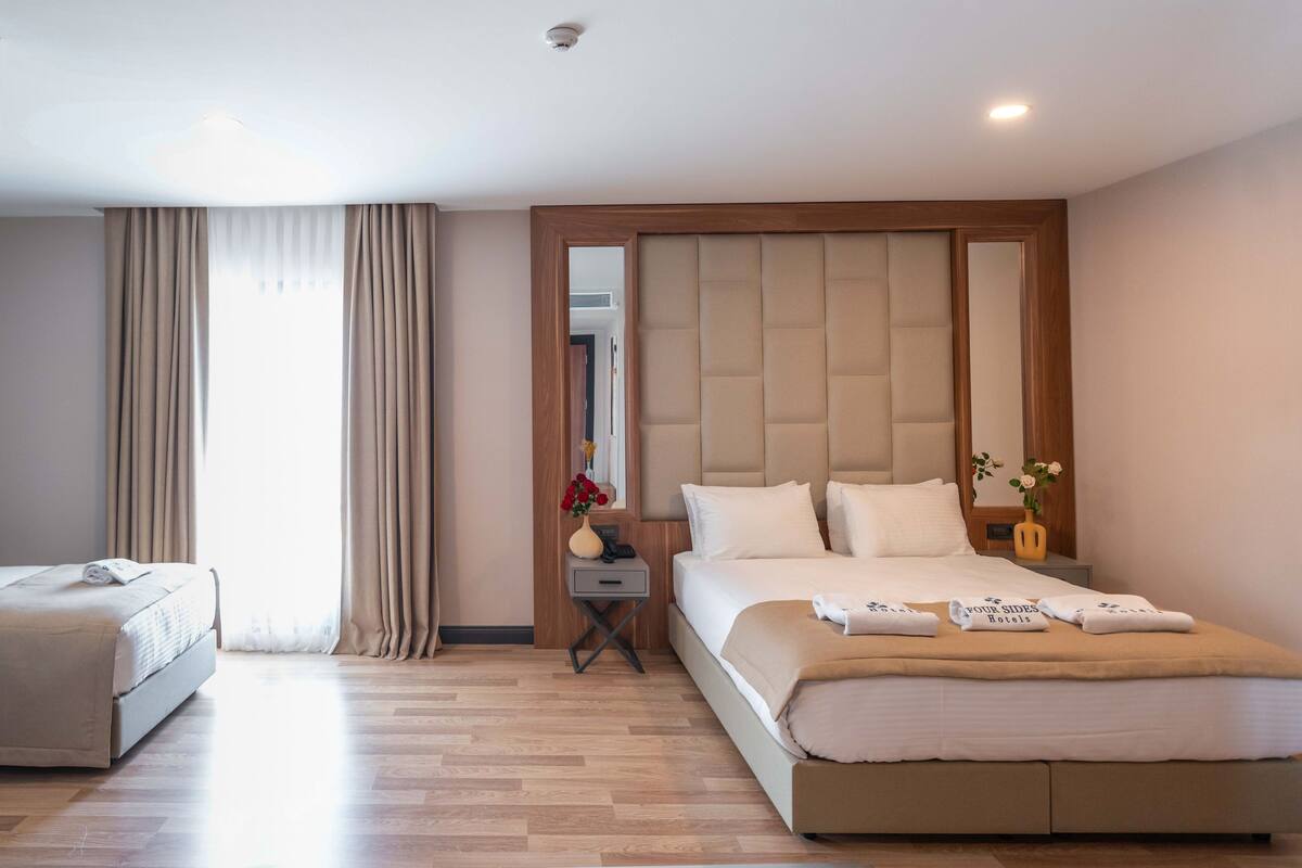 Quarto de hotel com uma cama grande, uma mesinha de cabeceira com um vaso de flores, um espelho e uma janela com cortinas.