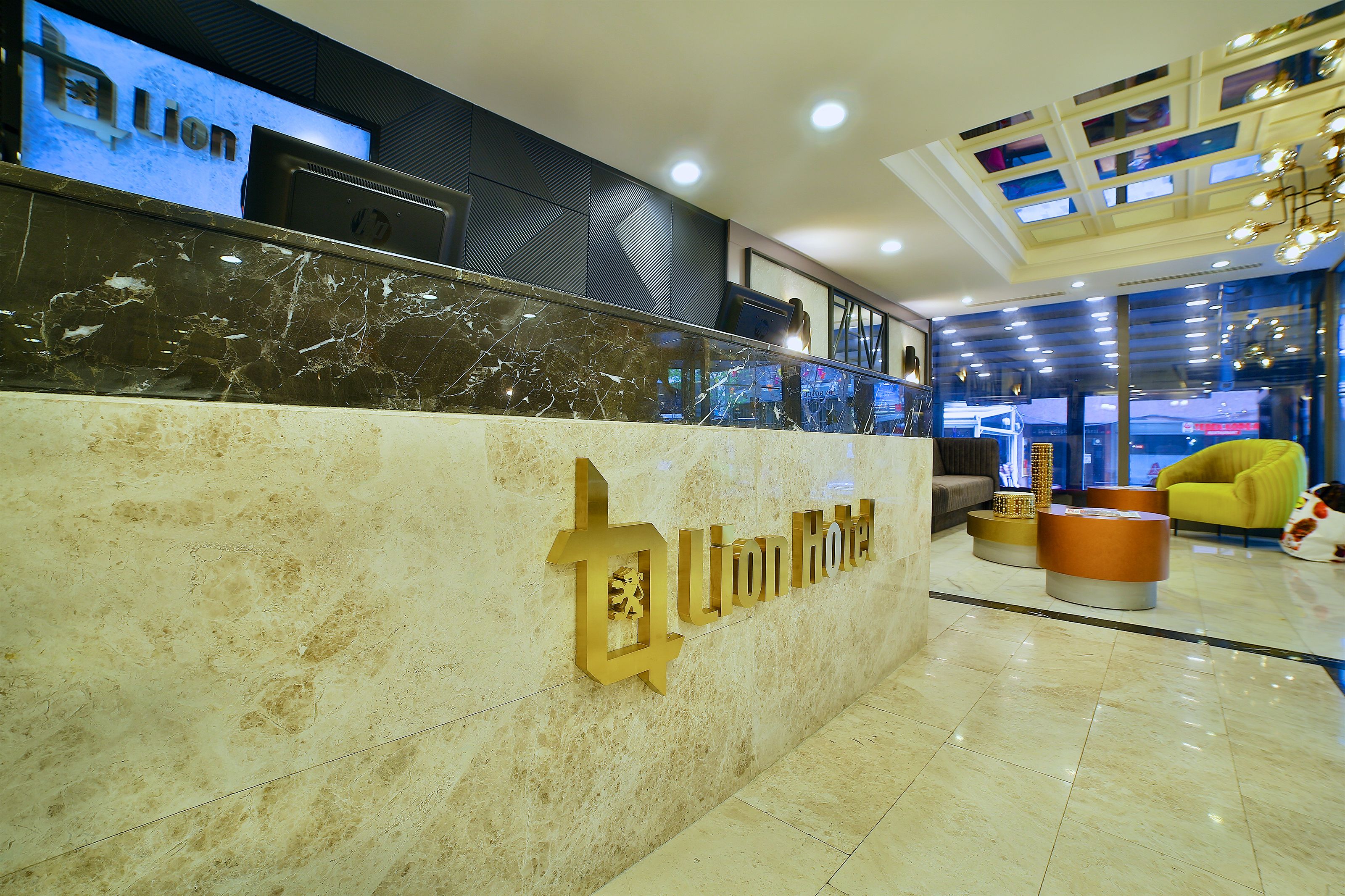 A hotel reception area with a marble counter, a digital screen displaying the hotel name, and a modern interior design with a yellow armchair and round ottoman.