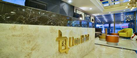 A hotel reception area with a marble counter, a digital screen displaying the hotel name, and a modern interior design with a yellow armchair and round ottoman.