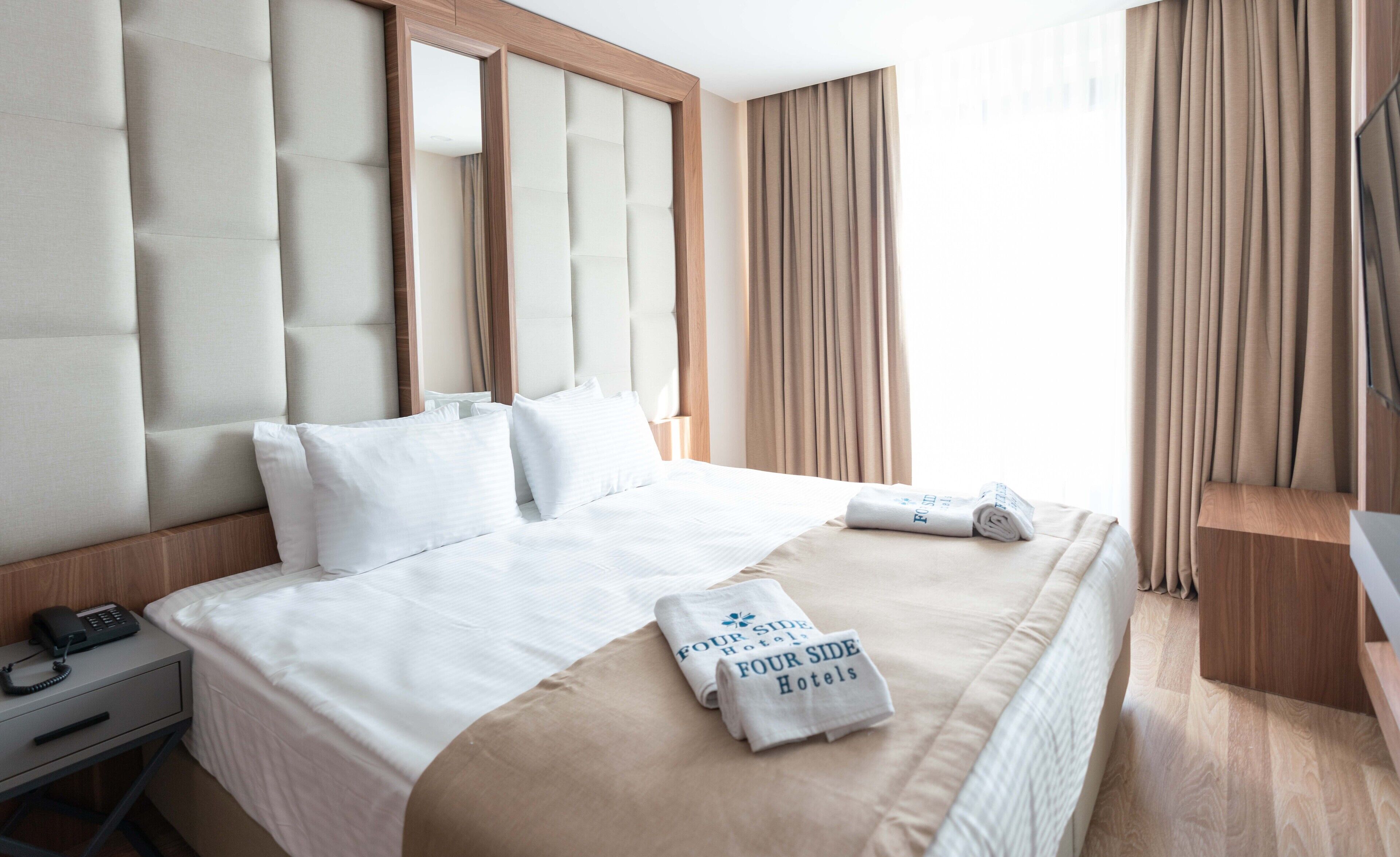 Hotellihuone, jossa on suuri sänky, sängyn vieressä puhelin ja hotellin nimellä varustettu, siististi taitettu pyyhe.