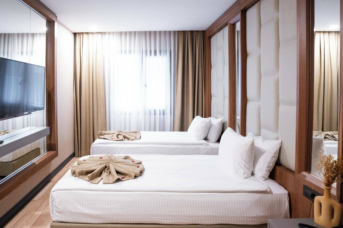 Quarto de hotel com duas camas, televisão, escrivaninha com telefone e um vaso com flores secas.