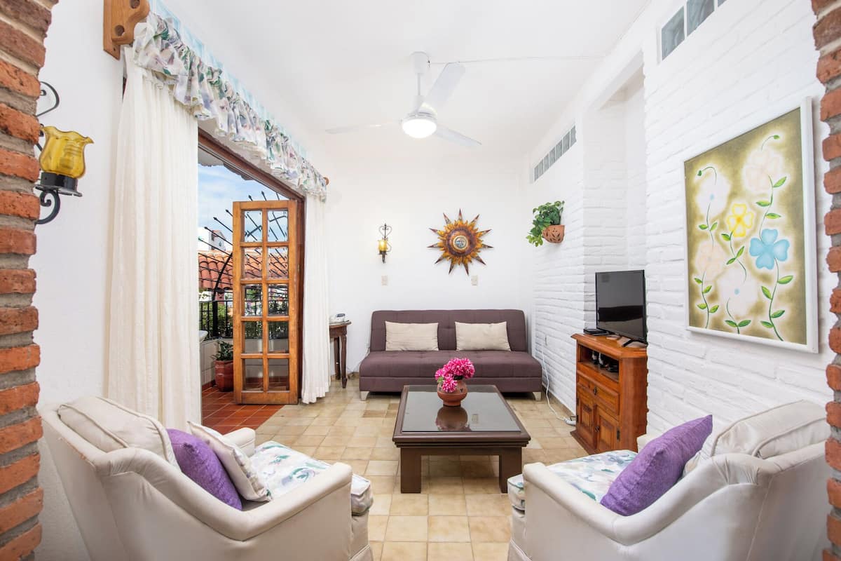 A living room with a sofa, two armchairs, a coffee table, a TV on a wooden stand, a decorative sun, and a painting on the wall.