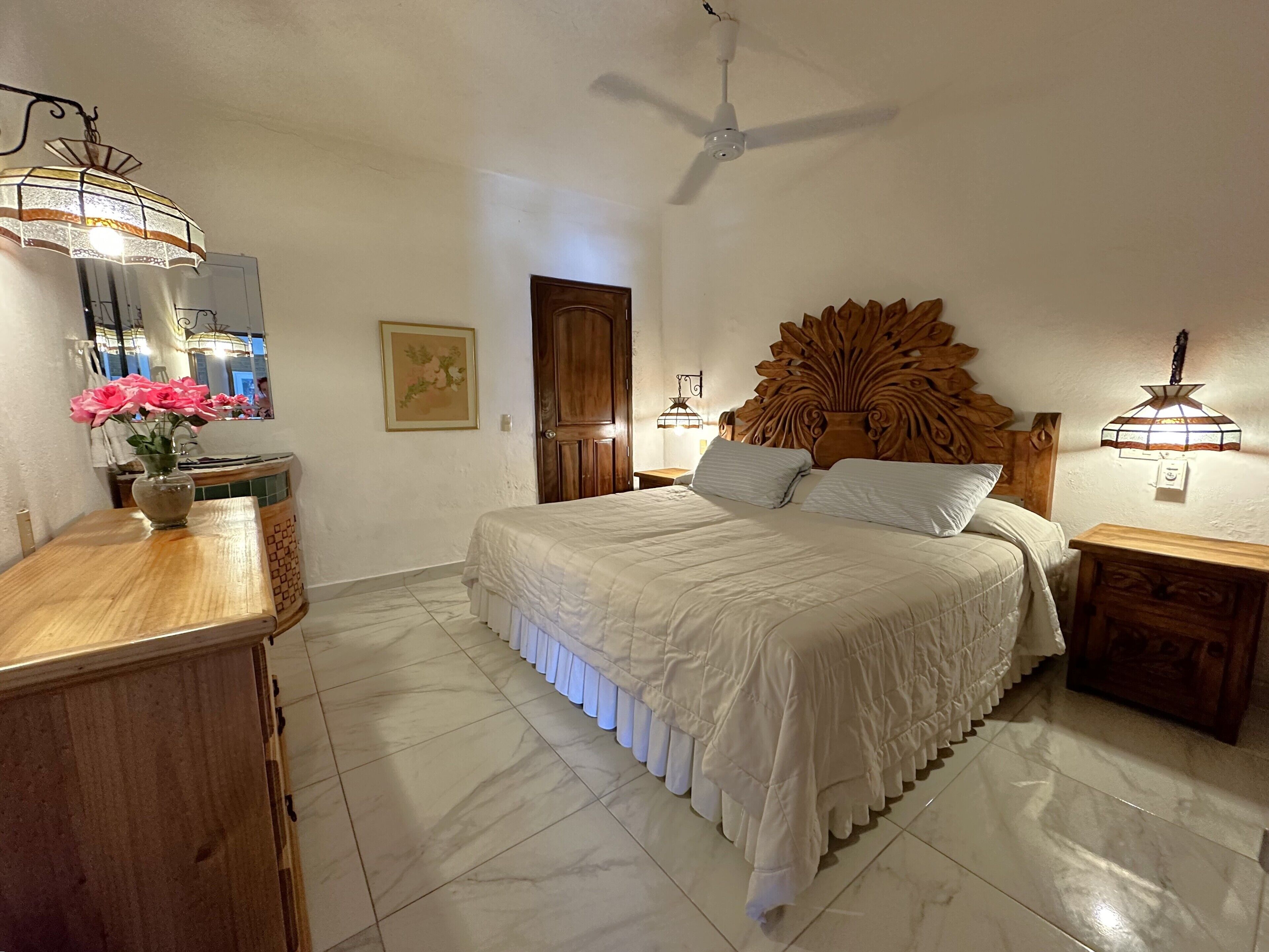 A bedroom with a large bed, a wooden headboard, a ceiling fan, and a nightstand with a lamp.