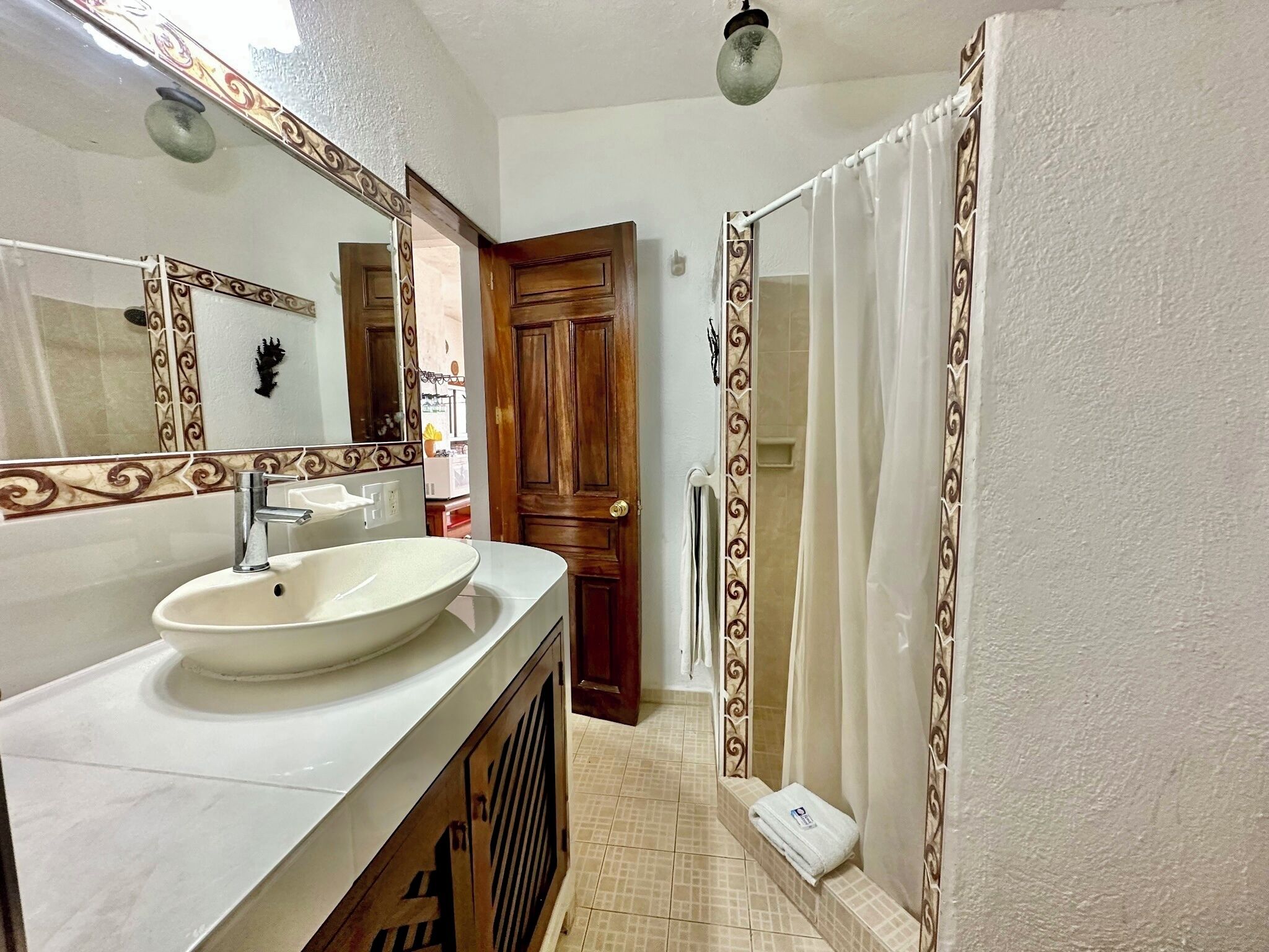 A bathroom with a large mirror, a sink, a shower with a curtain, and a door leading to another room.