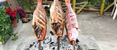 Tres pescados a la parrilla ensartados en brochetas, sobre carbón, con una fuente de mantequilla derretida cerca.