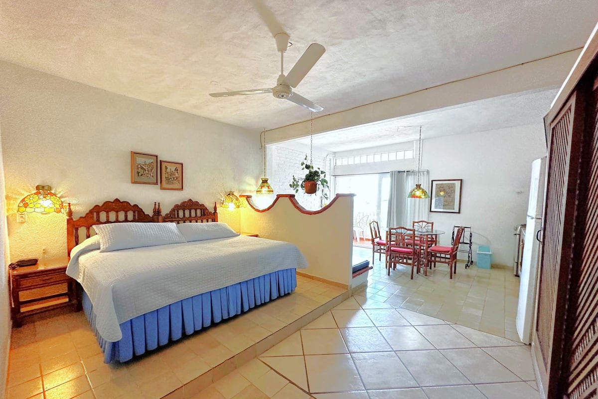 A bedroom with a bed, bedside lamps, a ceiling fan, and a dining area with chairs and a table.