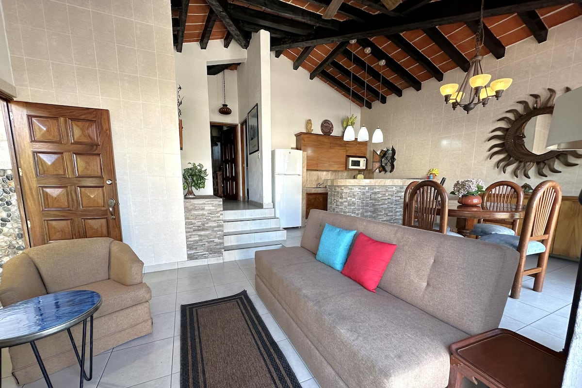 A cozy living room with a sofa, a small table, and a dining area with chairs.