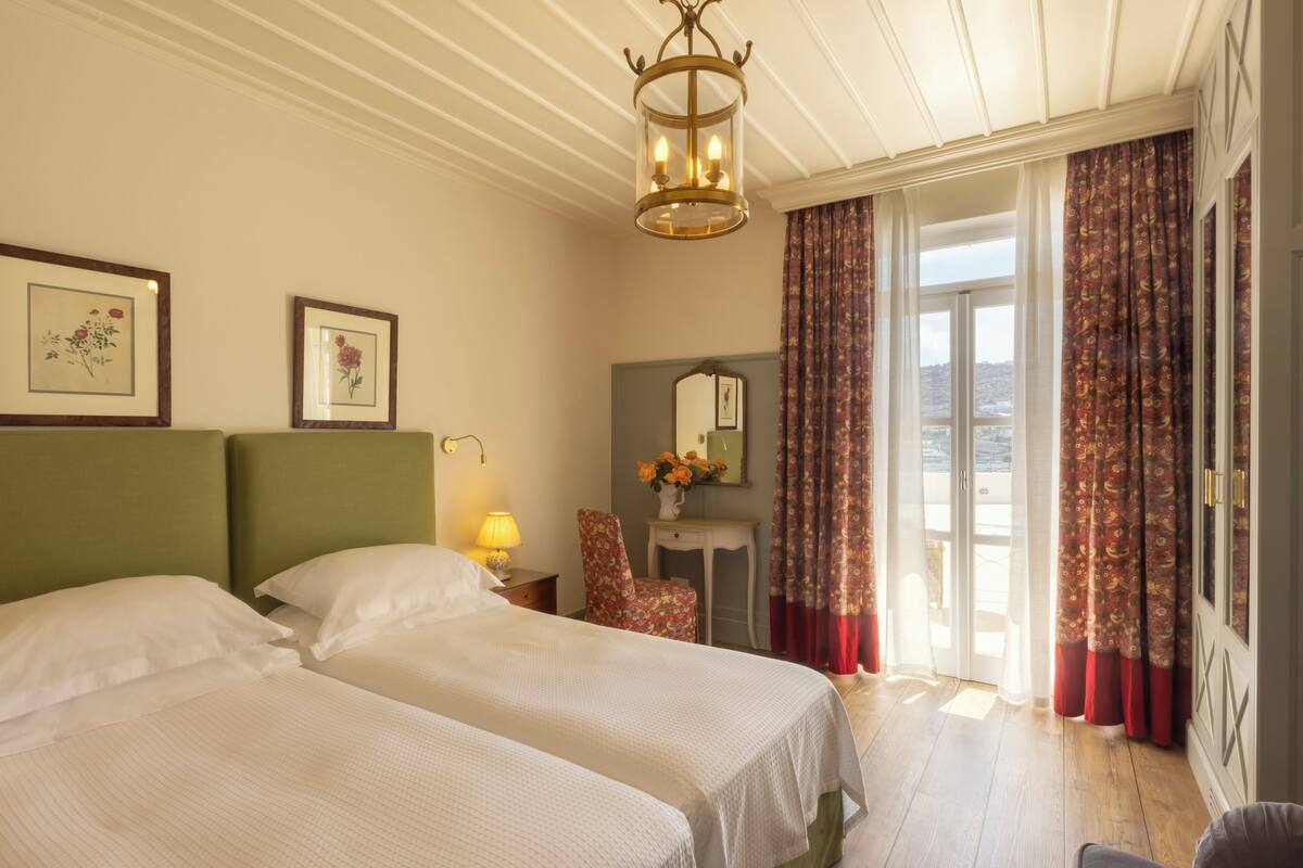 A hotel room with two beds, a nightstand with a lamp, a chair, a desk with a mirror, and a view of the outdoors through a door.