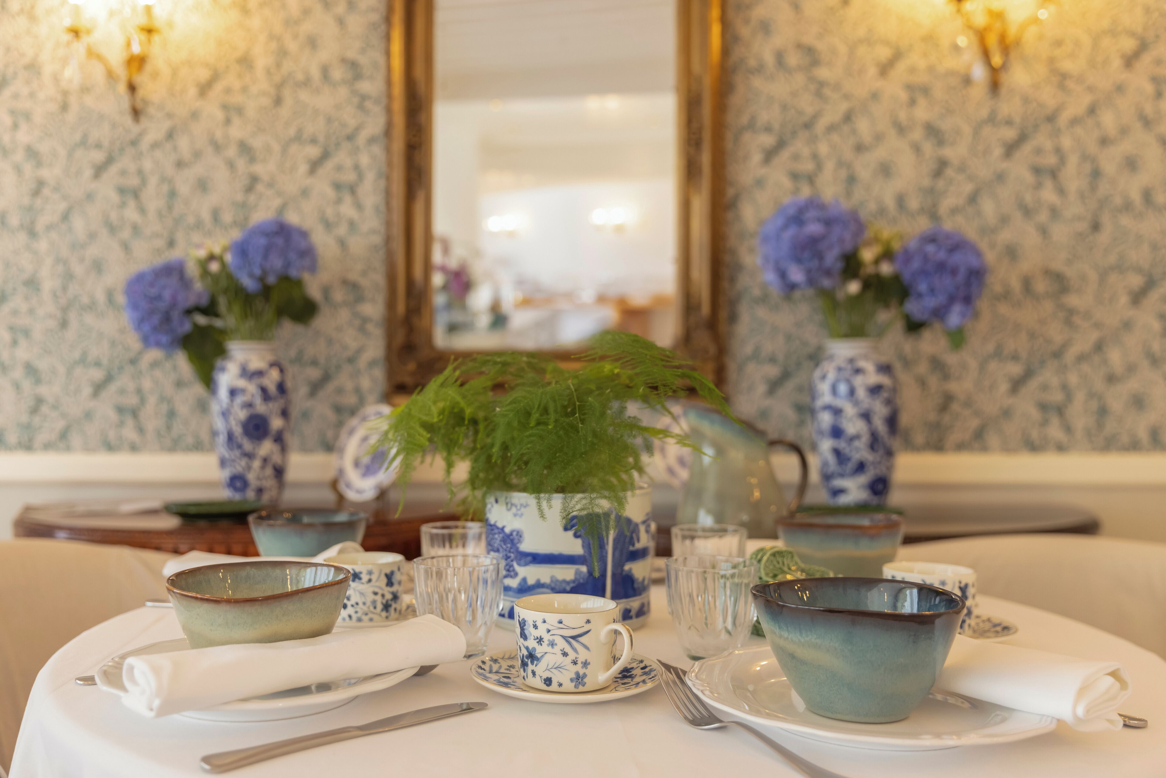 Une table à manger dressée avec de la vaisselle bleue et blanche à motifs, comprenant des bols, des tasses et un vase avec une plante verte.