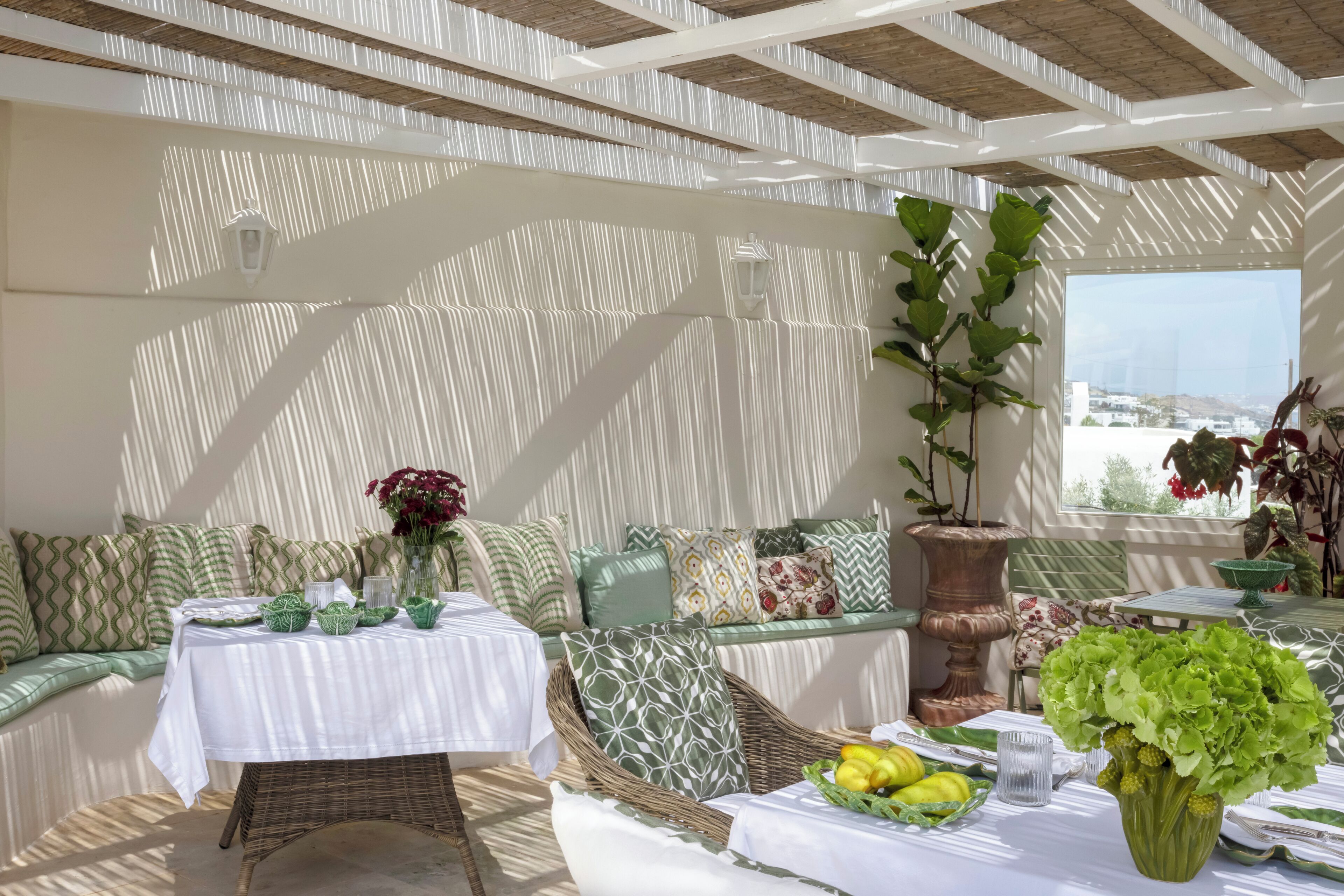 A sunlit dining area with a table set for two, a wicker sofa with patterned cushions, and a potted plant.
