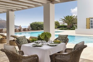 A covered outdoor dining area with wicker furniture, a pool, and a view of the sea. - Dorion Hotel (Mykonos)