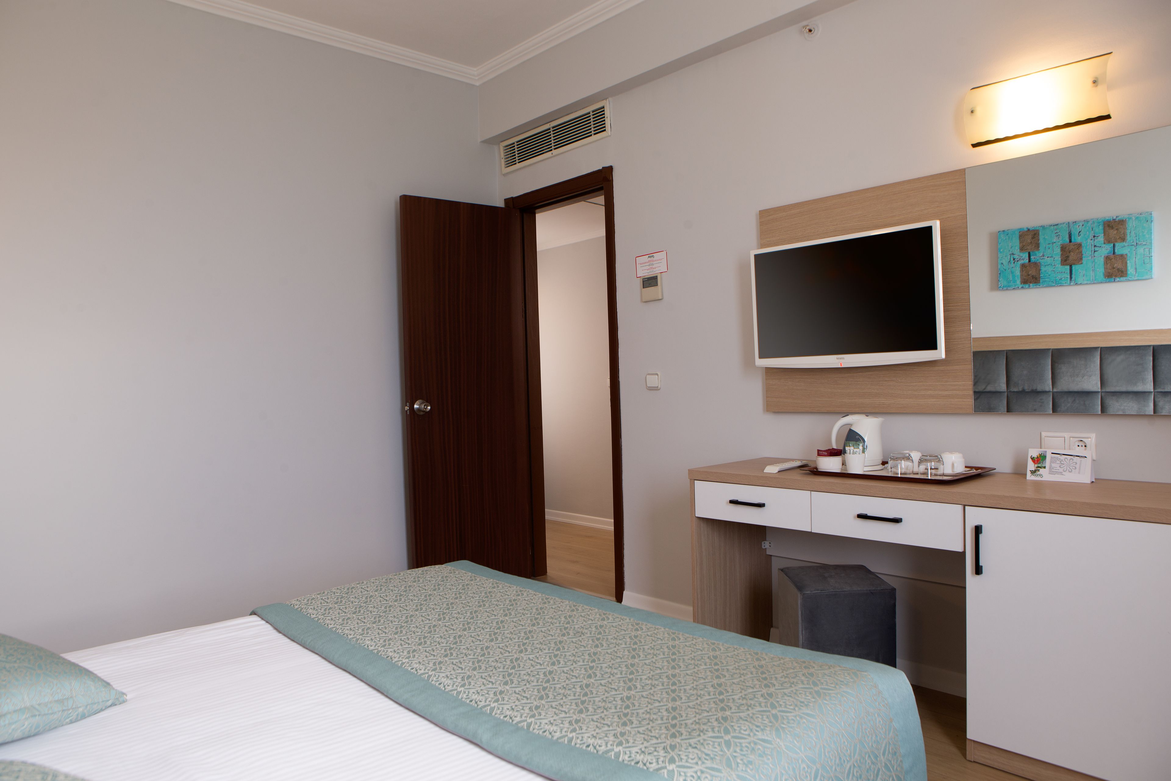 A hotel room with a bed, a desk, a chair, a television, and a small table with a kettle and cups.