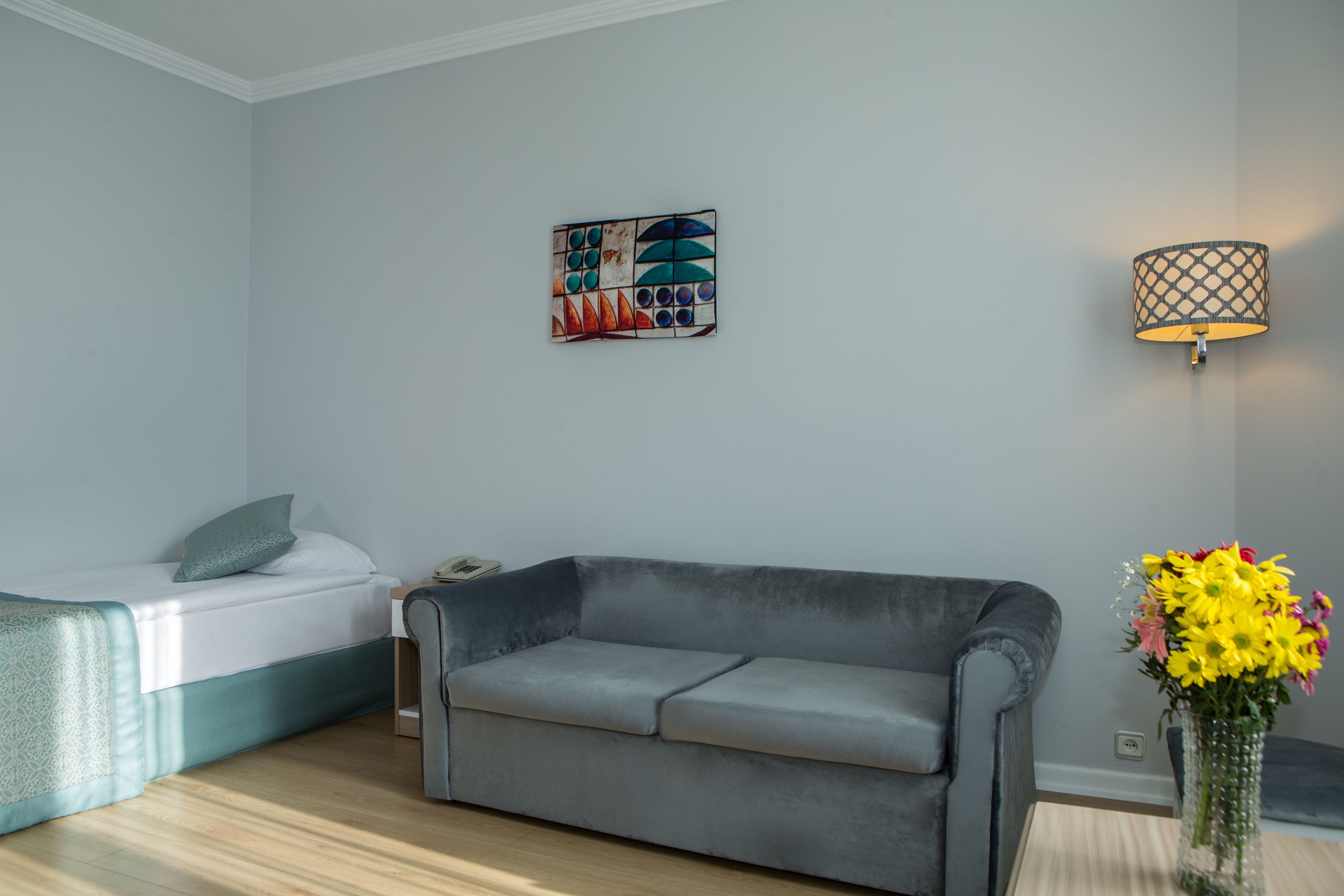 A modern living room with a grey sofa, a bed with a white duvet, a wall-mounted lamp, and a painting.