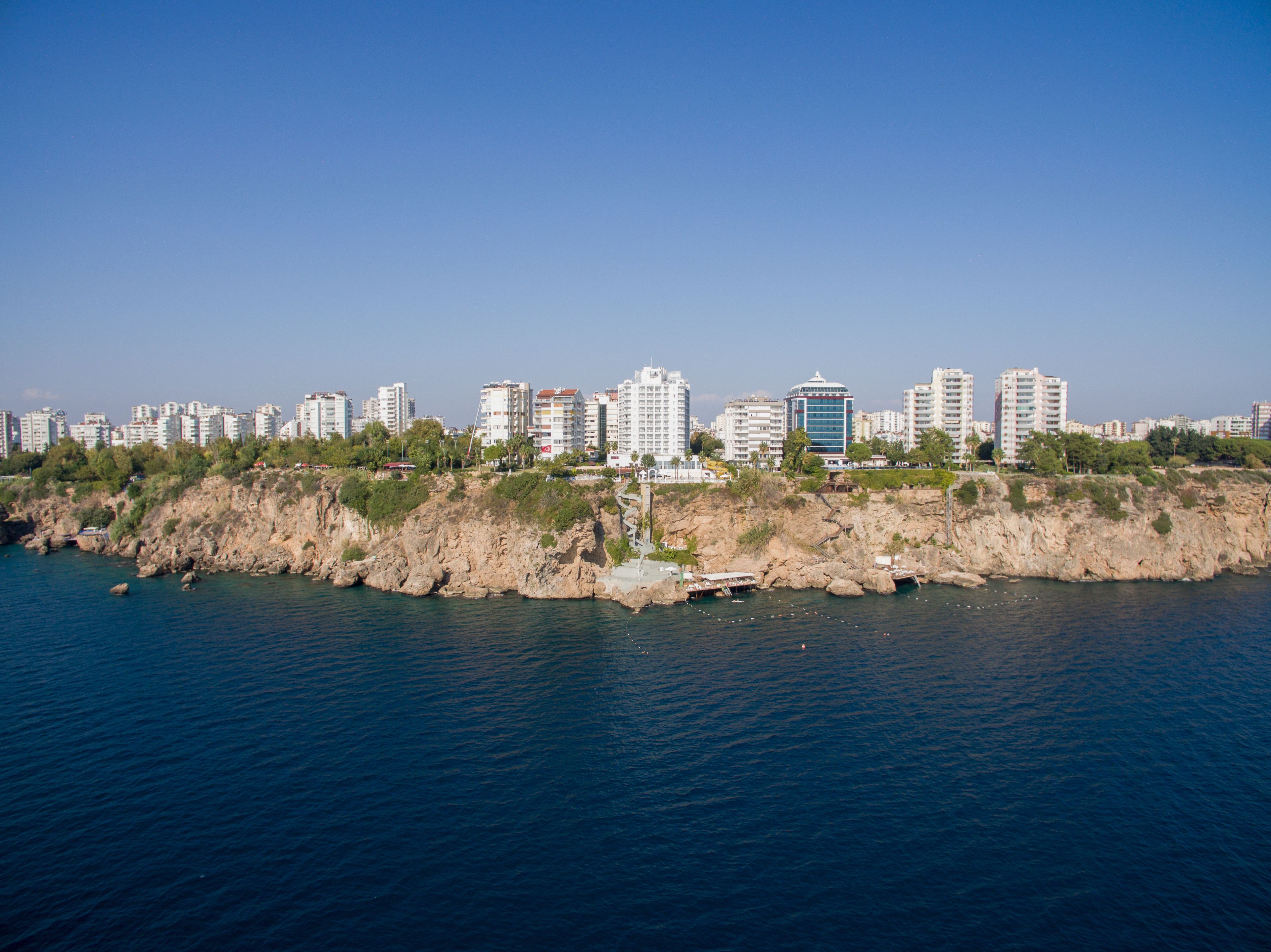A coastal cityscape with high-rise buildings, rocky cliffs, and a clear blue sky.