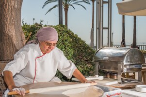 A chef preparing food outdoors with a portable grill and rolling pin, surrounded by greenery and palm trees.