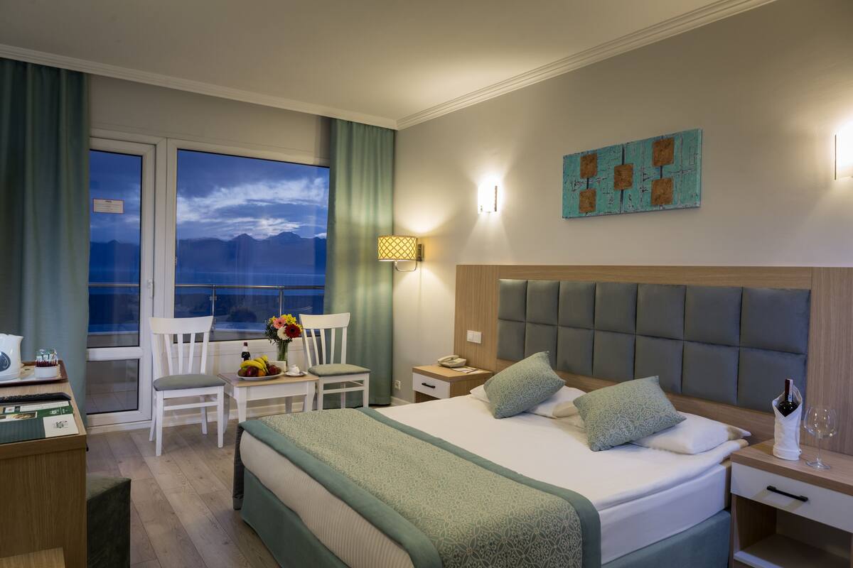 A hotel room with a large bed, a desk with a chair, a window with a view of mountains, and a wall-mounted artwork.