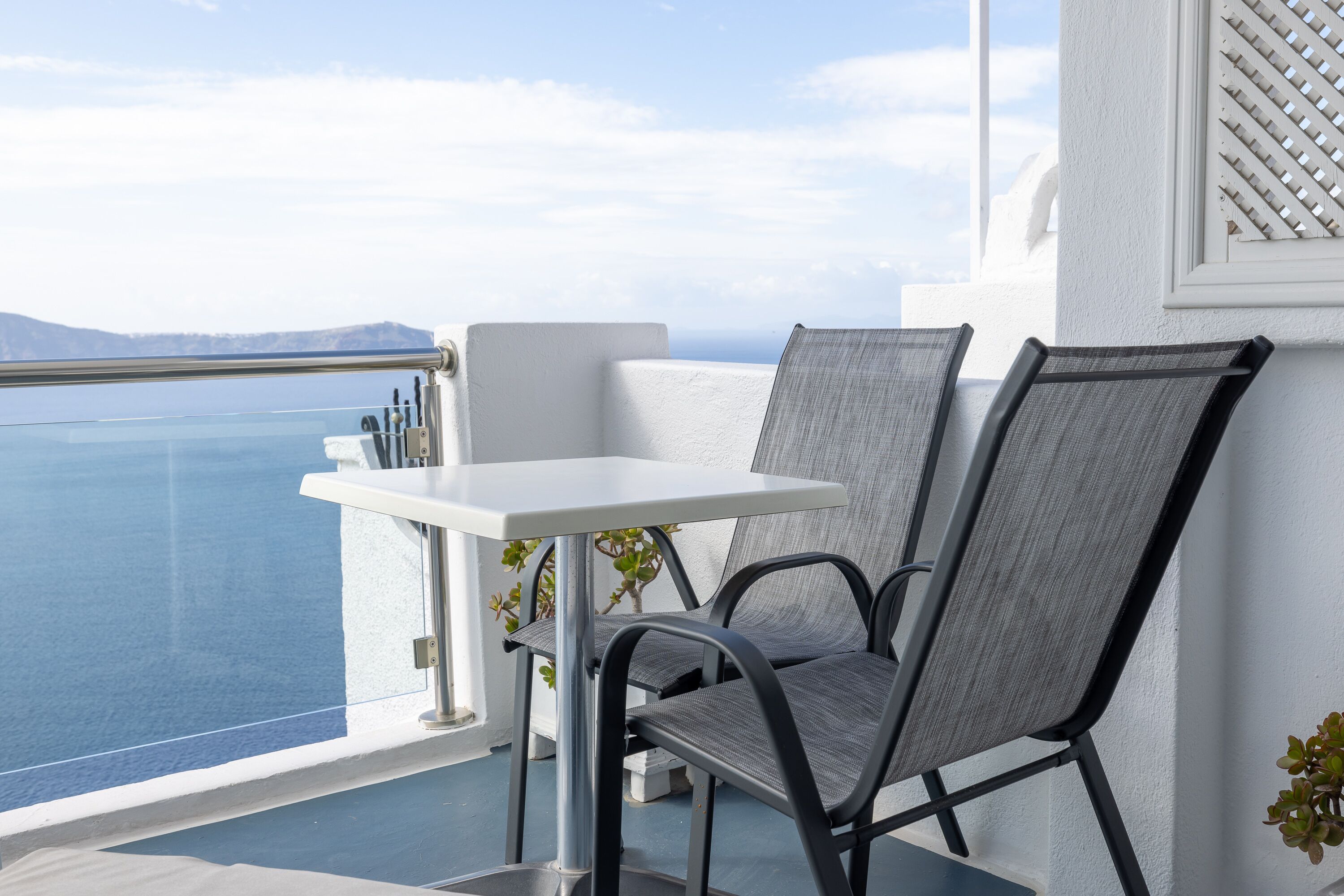 A balcony with a white table and two chairs, overlooking a clear blue sea and distant mountains.