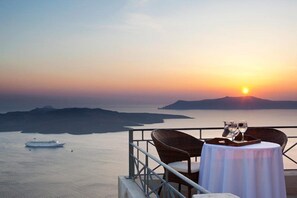 A table with two glasses and a tray, overlooking a sunset over water with a boat in the distance. - Asteras Villas (Santorini)
