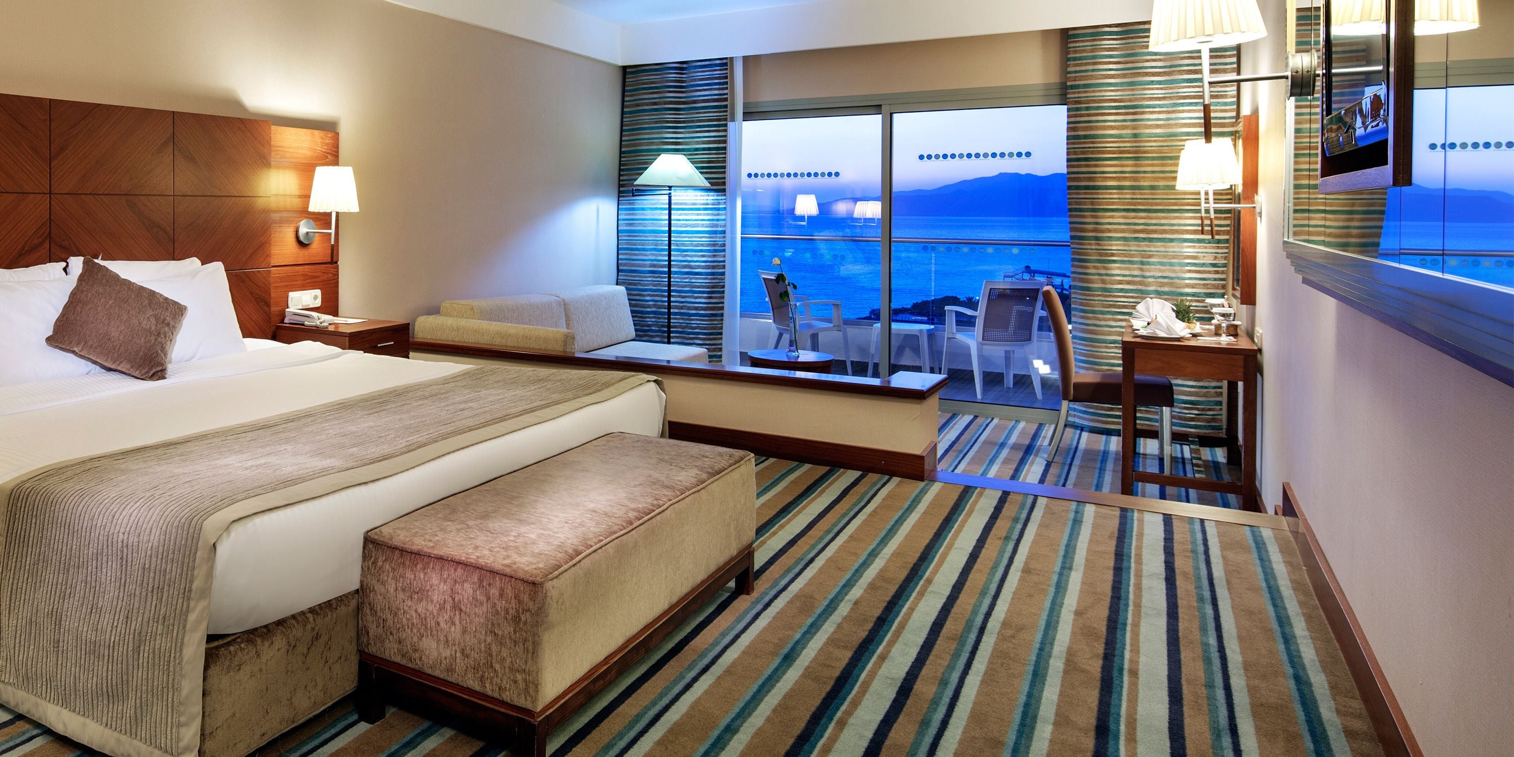 Habitación de hotel con una cama grande, una zona de estar con vista al mar, una pequeña mesa con sillas y un escritorio con espejo.
