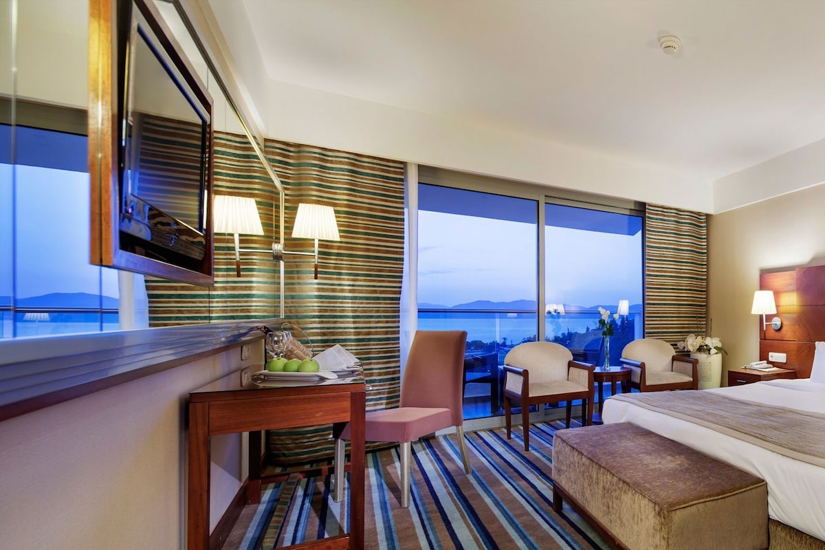 A hotel room with a large bed, a desk with a chair, a small table with a chair, and a view of the sea.