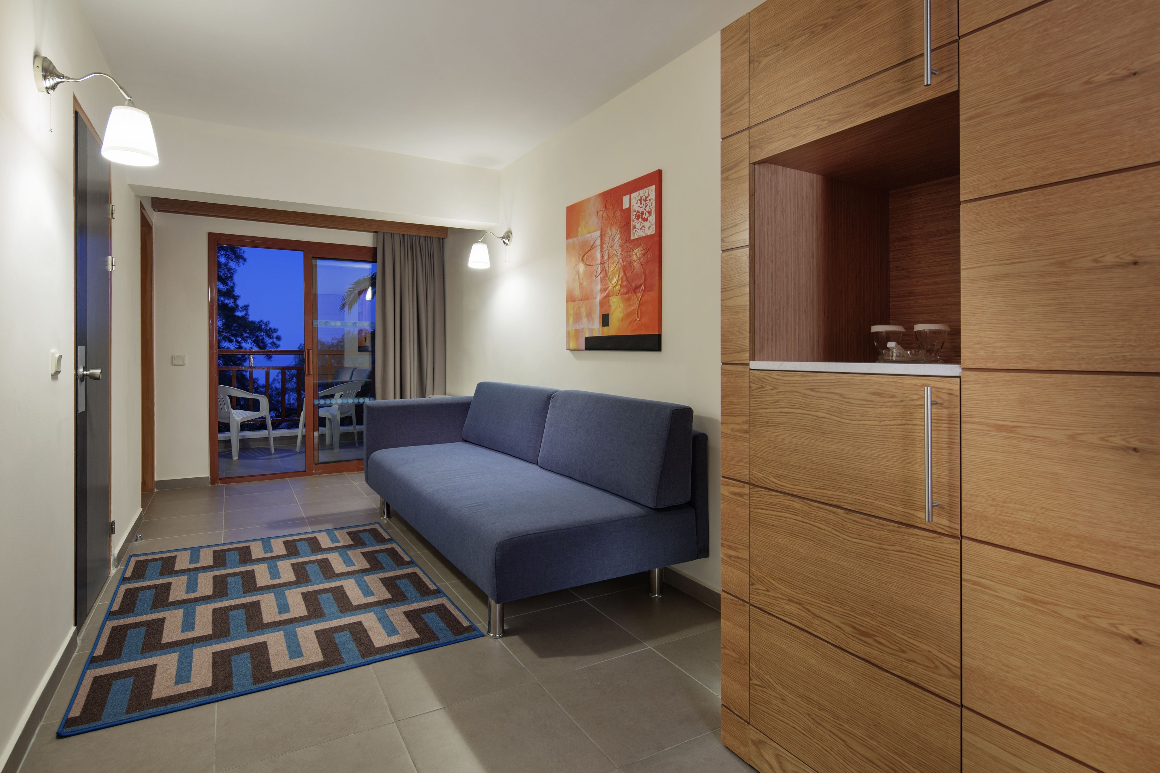 A modern living room with a blue sofa, wooden cabinets, a patterned rug, and a view through a glass door.
