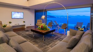 A modern living room with a large sofa, a coffee table with flowers, and a TV on the wall. There is a balcony with chairs and a view of the sea. - Pine Bay Holiday Resort (Kusadasi)