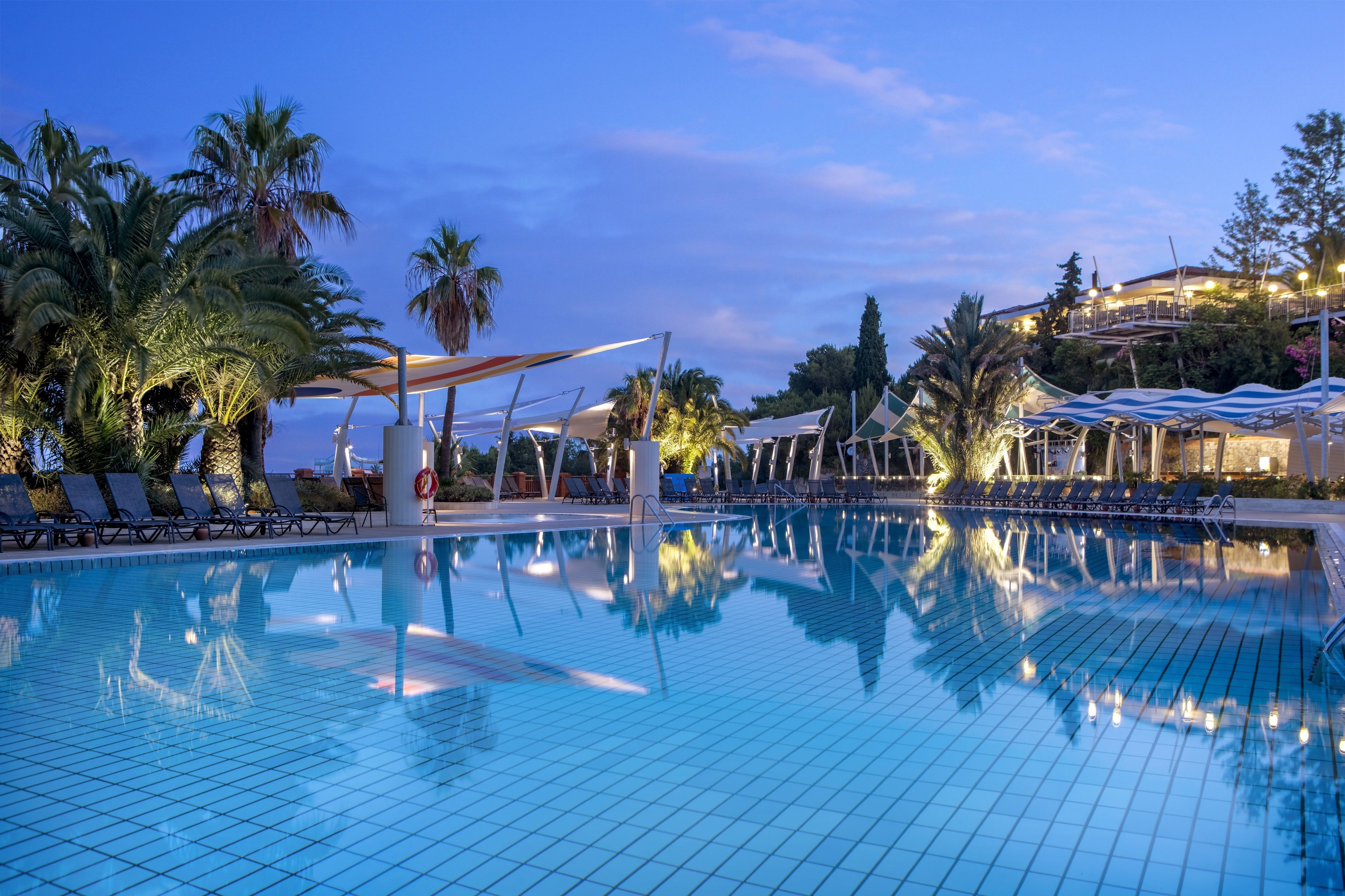 A swimming pool with lounge chairs and umbrellas, surrounded by palm trees and a building in the background.