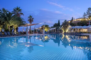 Una piscina con sillas de descanso y sombrillas, rodeada de palmeras y un edificio al fondo.
