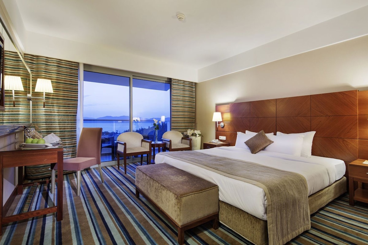 A hotel room with a large bed, a desk, two chairs, a small table, a bench, a nightstand, a lamp, and a view of the city.