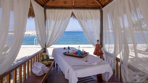 A beachside cabana with a wooden ceiling, white curtains, a massage table, and a tray of amenities. - Pine Bay Holiday Resort (Kusadasi)