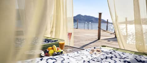 A beach view from a room with a bed, a table with fruits and a drink, and a view of the beach and mountains.