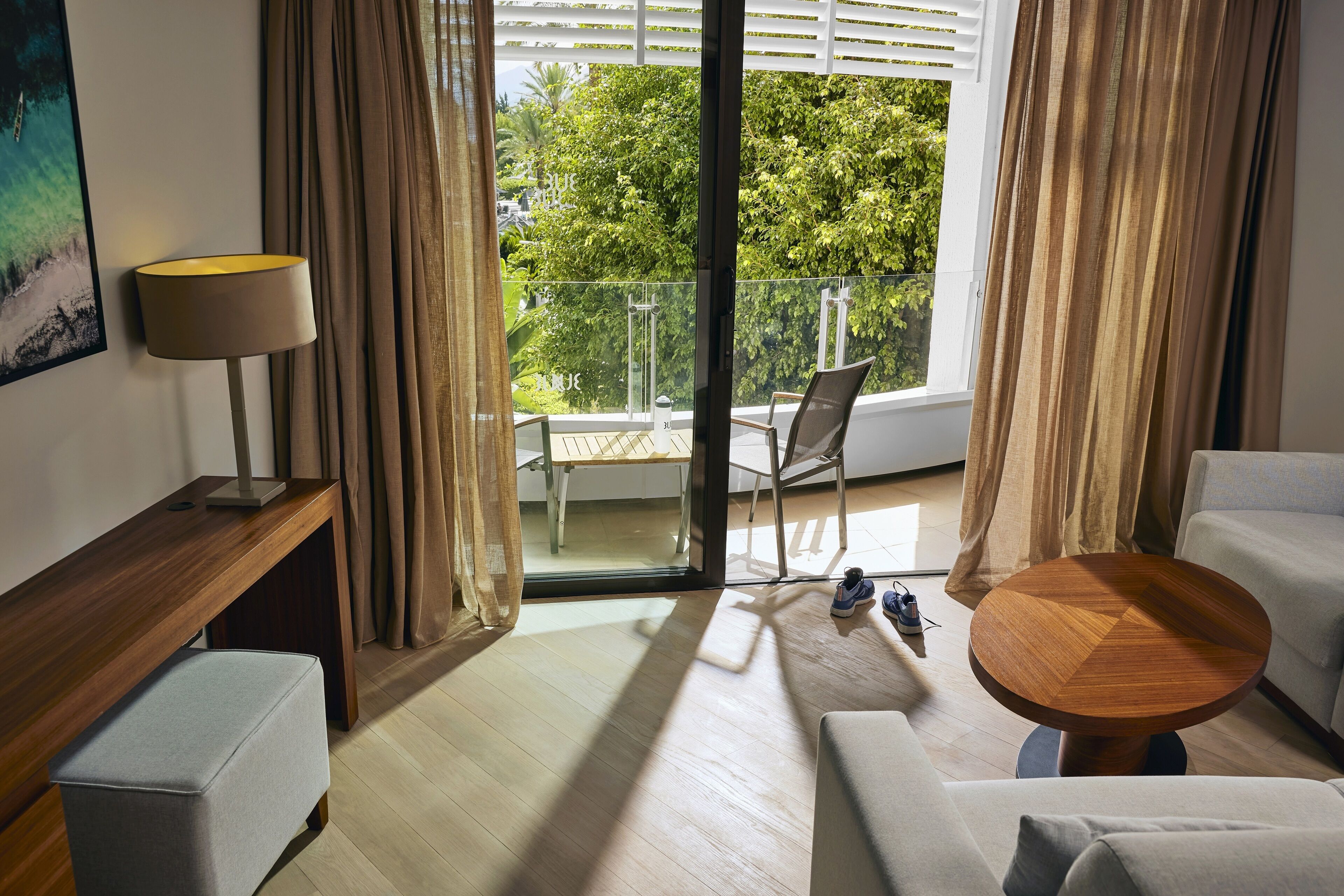 A modern hotel room with a balcony, a wooden side table, a round wooden coffee table, a sofa, and a chair.