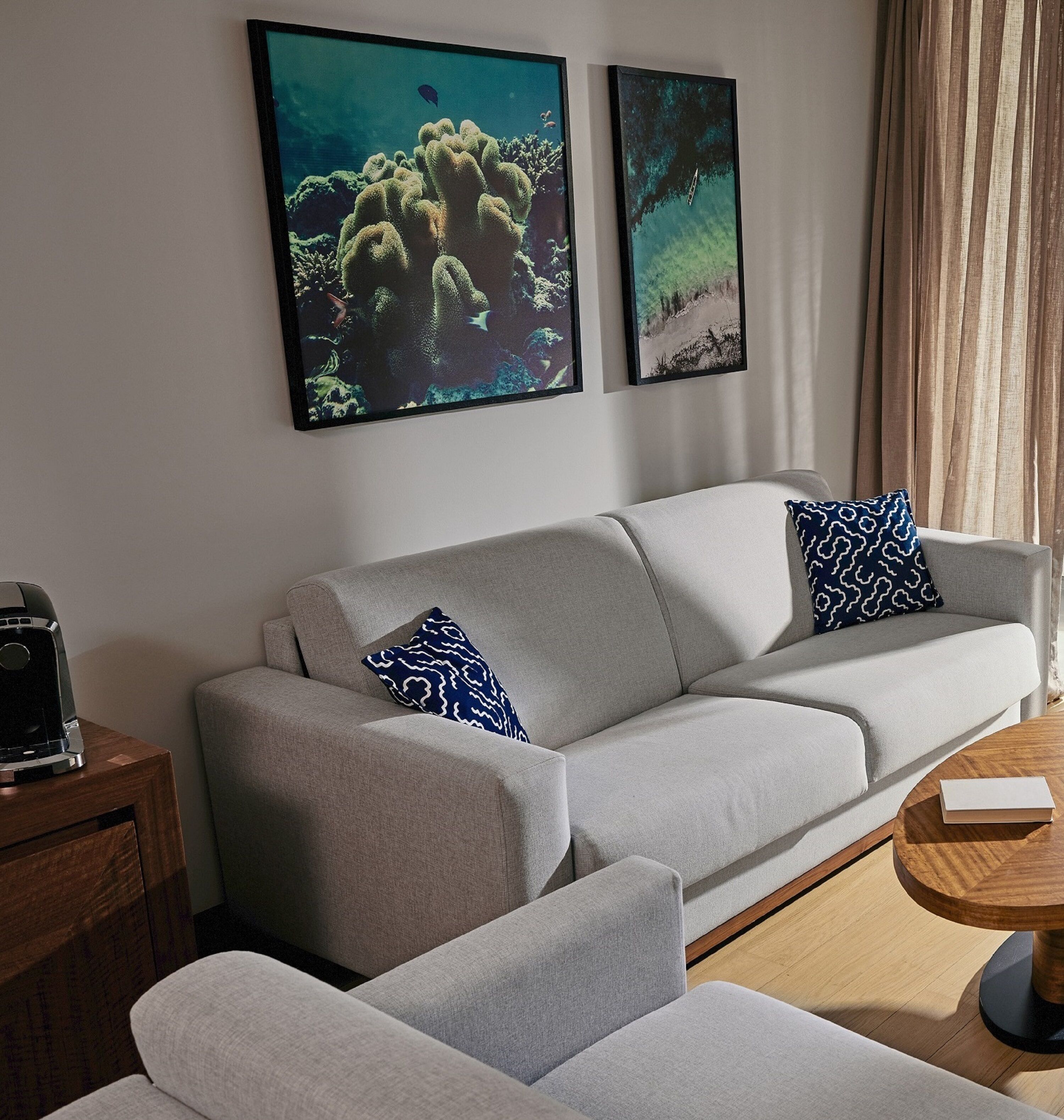 A modern living room with a grey sofa, two patterned pillows, a coffee machine on a wooden side table, and two framed artworks on the wall.