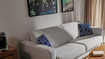 A modern living room with a grey sofa, two patterned pillows, a coffee machine on a wooden side table, and two framed artworks on the wall.