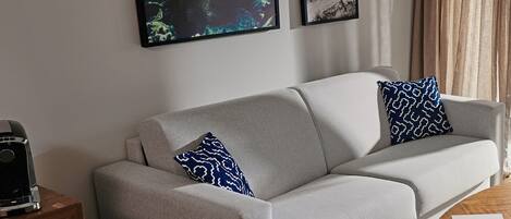 A modern living room with a grey sofa, two patterned pillows, a coffee machine on a wooden side table, and two framed artworks on the wall.