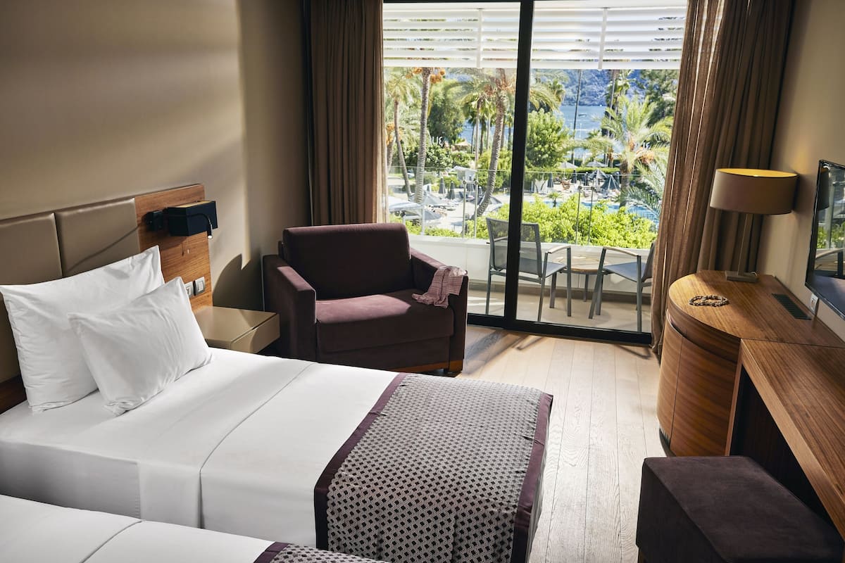 A hotel room with a bed, a chair, a desk, and a view of a pool and palm trees.