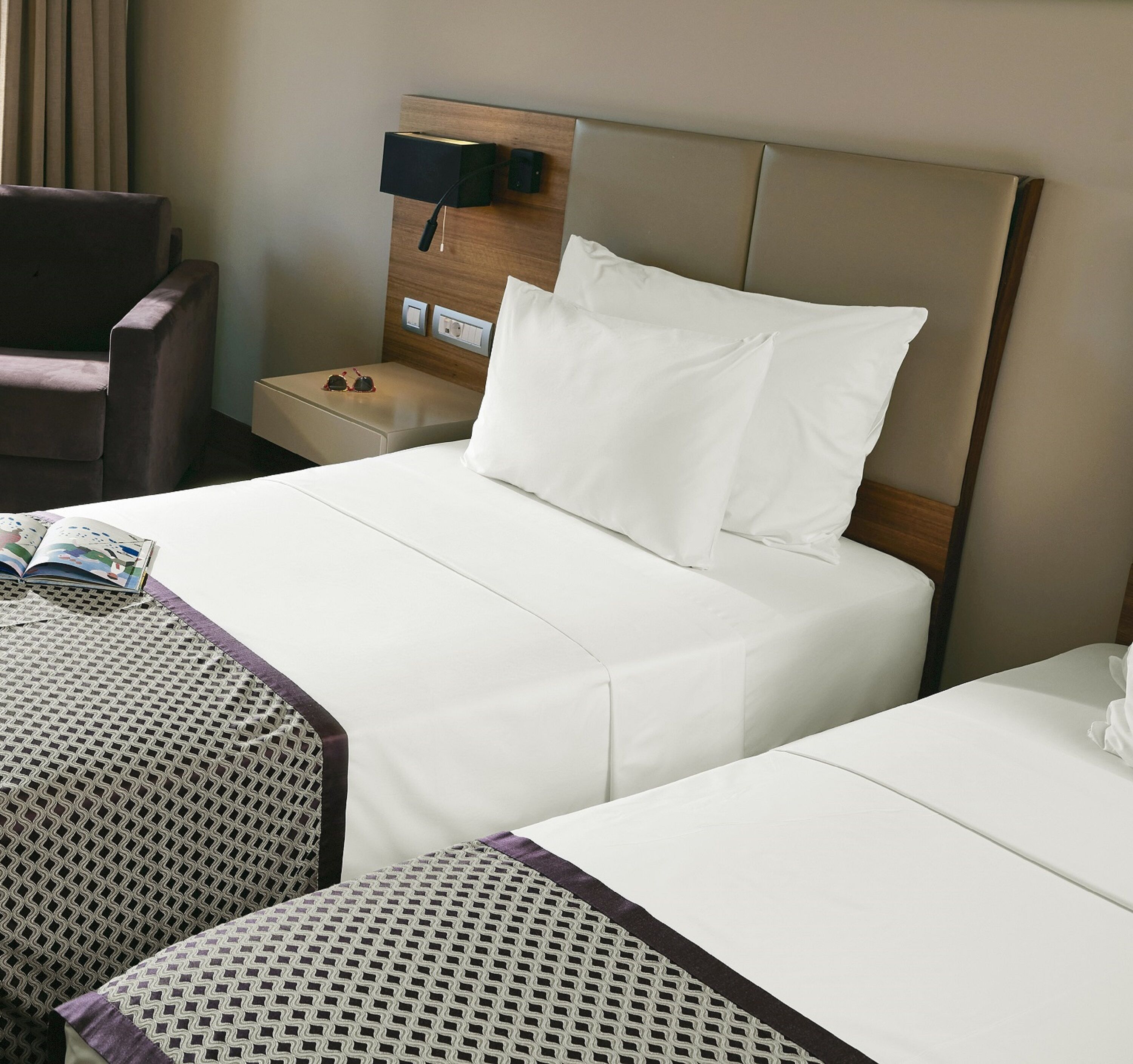 A hotel room with two beds, a nightstand, a chair, and a small table.