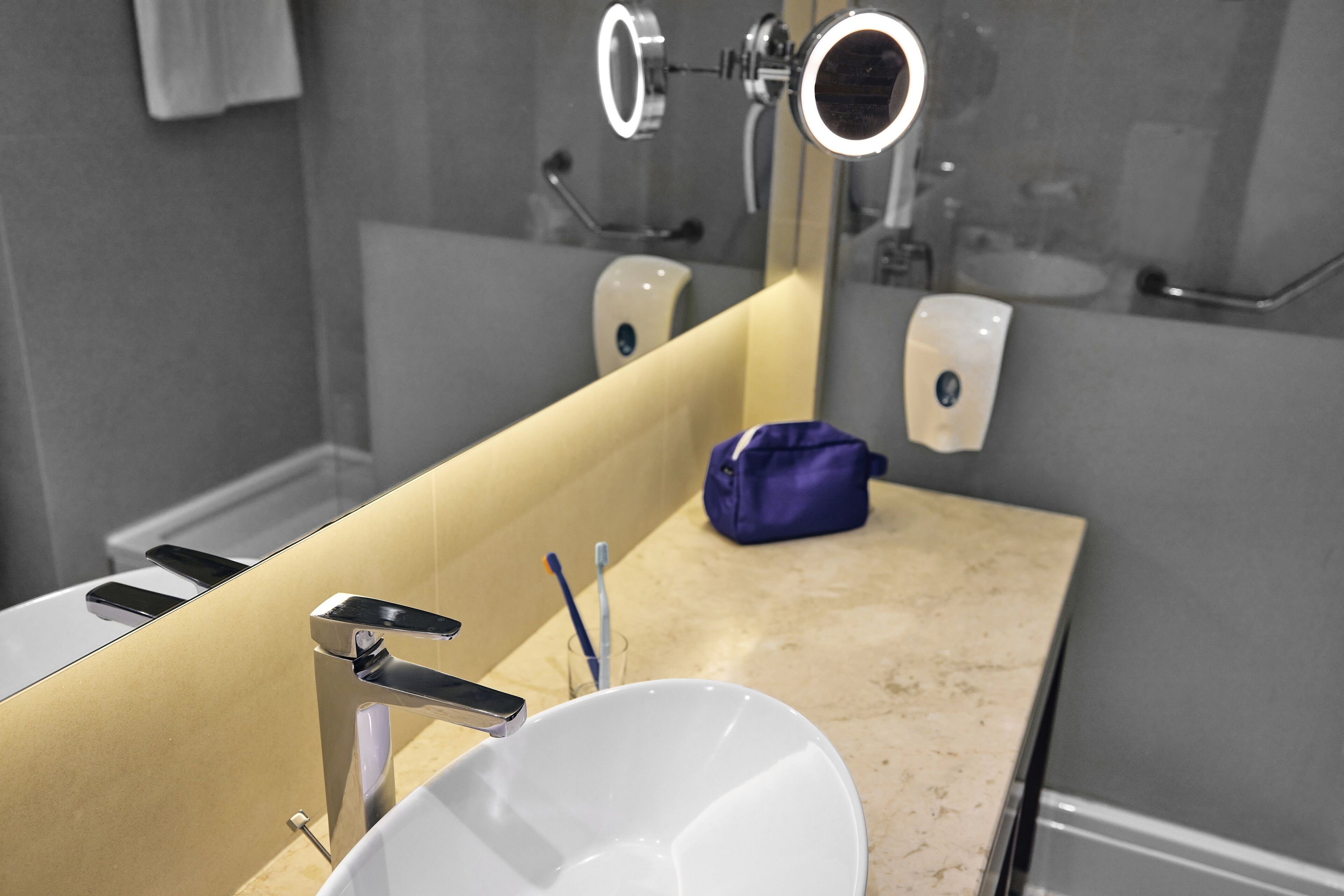 Ein Badezimmer mit einem weißen Waschbecken, einem Spiegel, einem Handspiegel, einer Seifenspender und einer blauen Tasche auf der Ablagefläche.