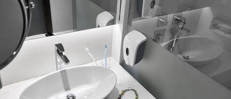 Un baño con un lavabo blanco, un espejo, un cabezal de ducha manual y un dispensador de jabón.