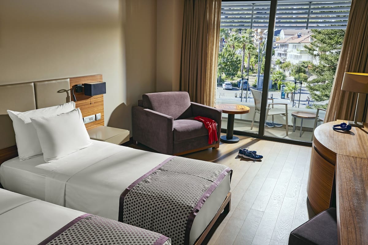 A hotel room with two beds, a seating area with a chair and a small table, and a view of the city through large windows.