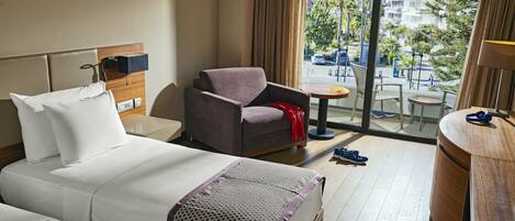 A hotel room with two beds, a seating area with a chair and a small table, and a view of the city through large windows.