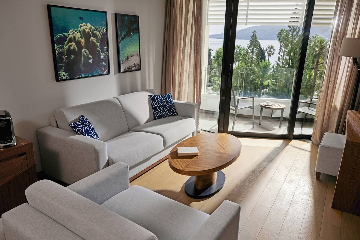 A modern living room with a sofa, a coffee table, and a balcony with a view of trees and a body of water.